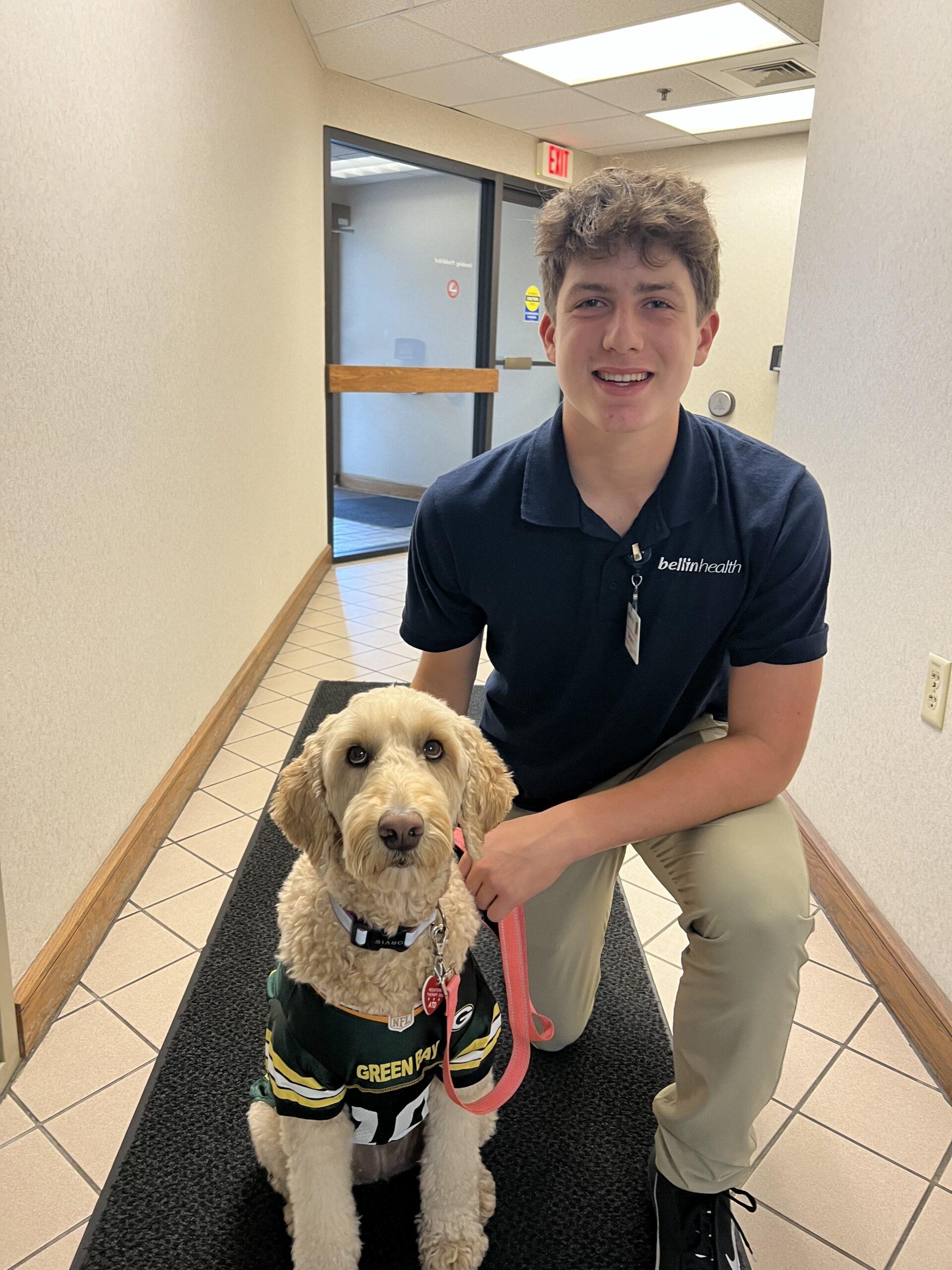 A young male kneels next to his golden colored dog smiling.