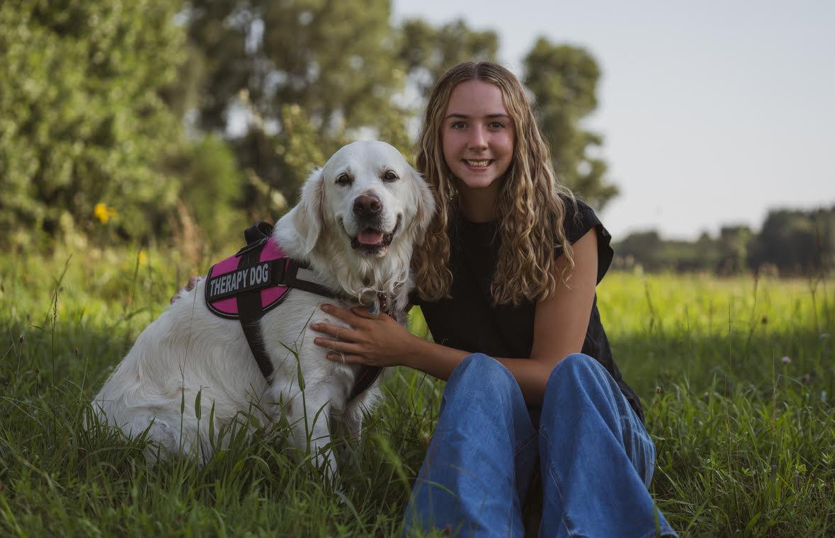 A young blonde haired junior handler sits next to her light colored golden retriever in a harness that says therapy dog.