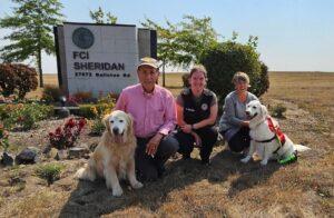 Two therapy dog handlers kneel with their dog with a woman between them in front of a FCI sign.