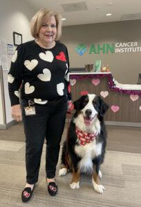 A woman in a heart sweater stands next to a tri colored therapy dog sitting next to her.